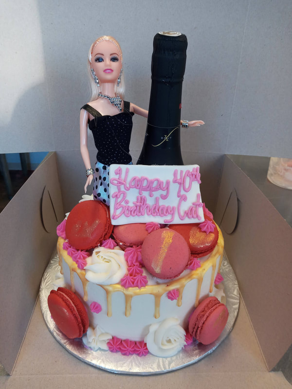White birthday cake with gold drip icing, pink macarons, the top of a wine bottle and a Barbie doll