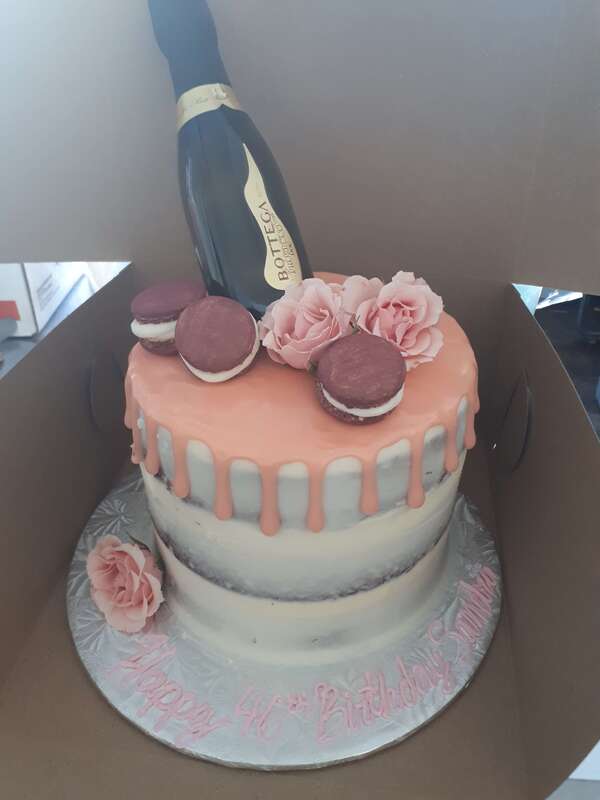 Lightly iced white cake witth drip icing, pink roses, macarons and a bottle of Bottega champagne